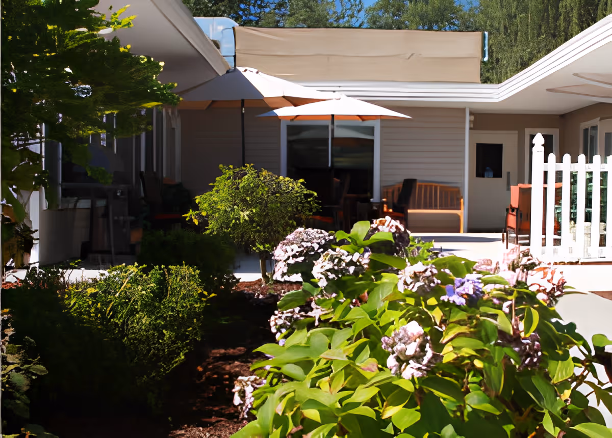 Flowering bushes in the foreground with a shaded patio area, umbrellas, benches and the building facade in the background.