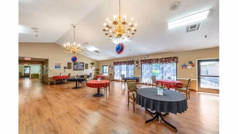Spacious dining/activity room with round tables covered in tablecloths, chandeliers, and large windows letting in natural light.