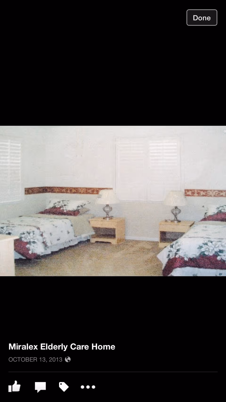A bedroom with two single beds placed against adjacent walls, each bed covered with floral patterned bedding. Between the beds are two wooden nightstands, each with a decorative lamp. The room has light-colored walls with a floral border and windows with white shutters.