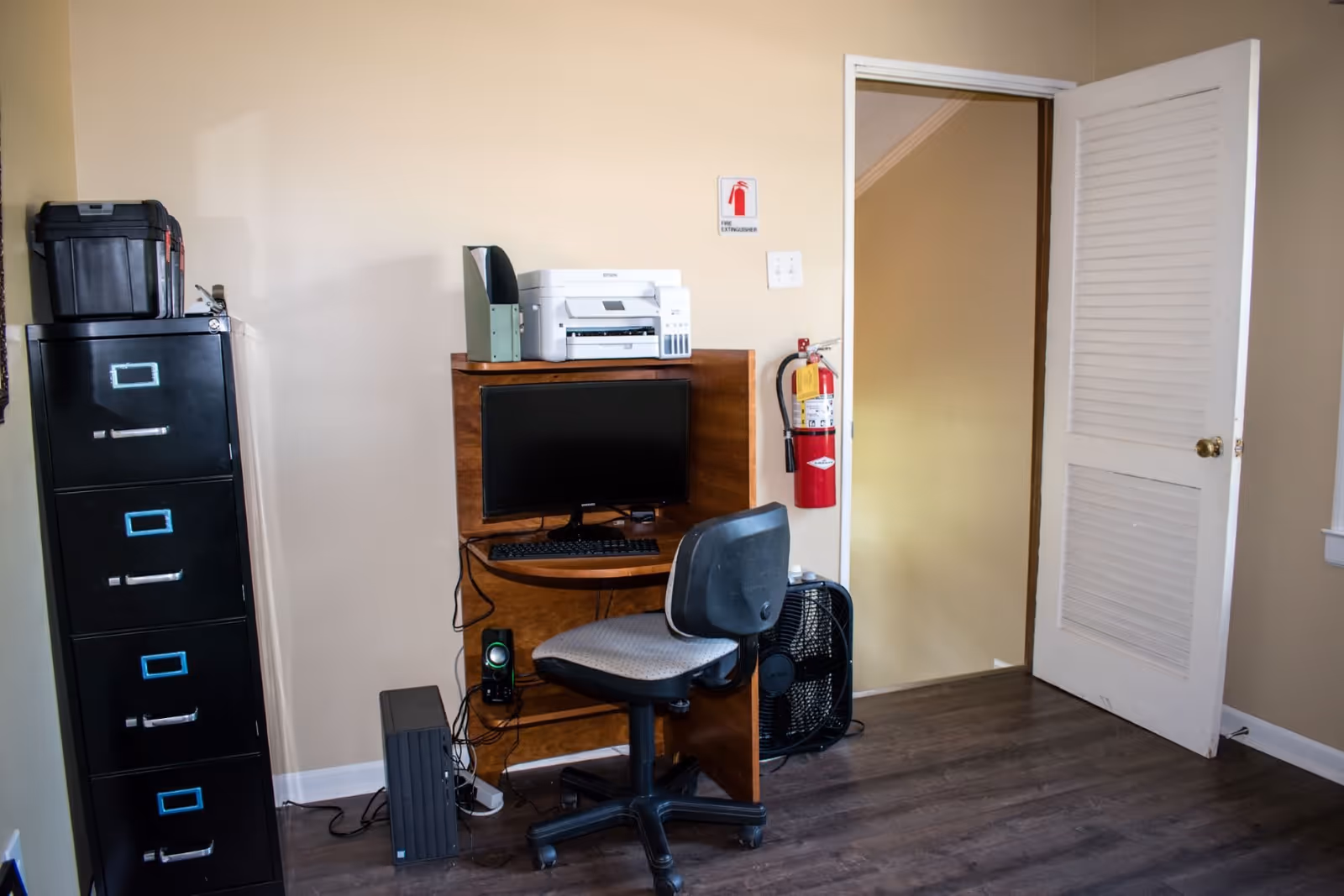 A small office corner with a wooden desk holding a computer monitor, keyboard, printer, and file organizer. A black office chair is positioned in front of the desk. To the left is a tall black filing cabinet with a toolbox on top. A fire extinguisher is mounted on the wall near an open white door. The room has beige walls and dark wood flooring.