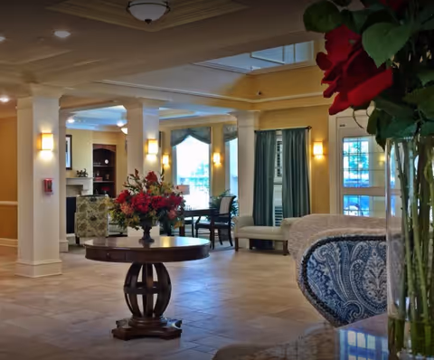 A spacious and well-lit living area in a senior living facility featuring a round wooden table with a floral arrangement in the center. The room has large windows with green curtains, comfortable seating including a patterned armchair and a bench, and decorative columns. Warm lighting fixtures are mounted on the walls and ceiling.