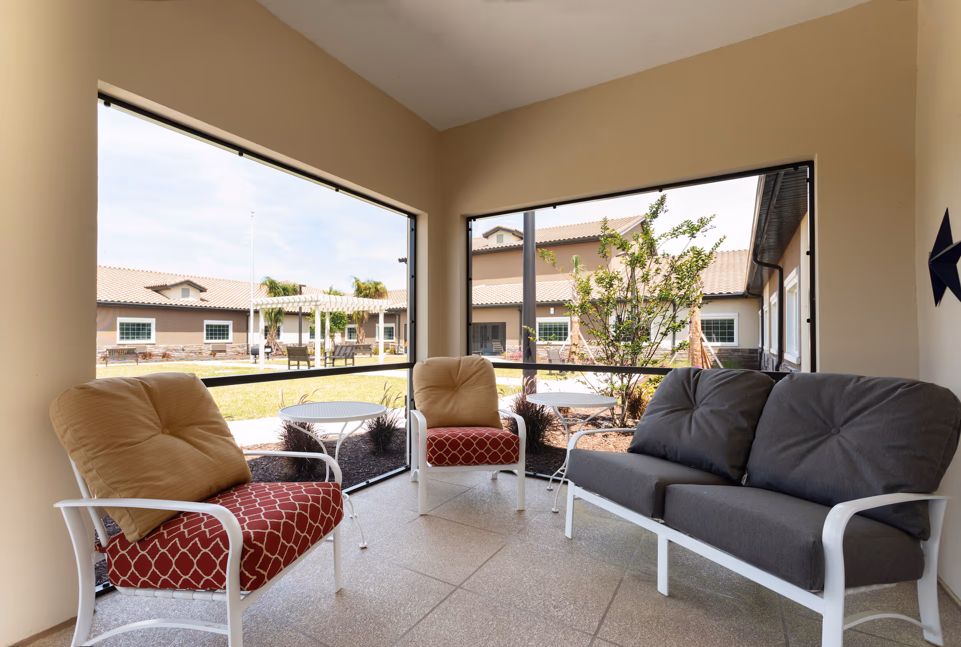 A screened-in porch area with cushioned outdoor seating including two armchairs with red patterned cushions and tan pillows, and a gray cushioned loveseat. Two small white round tables are placed between the chairs. Outside the porch, there is a landscaped courtyard with plants and a building with beige walls and a tiled roof in the background.