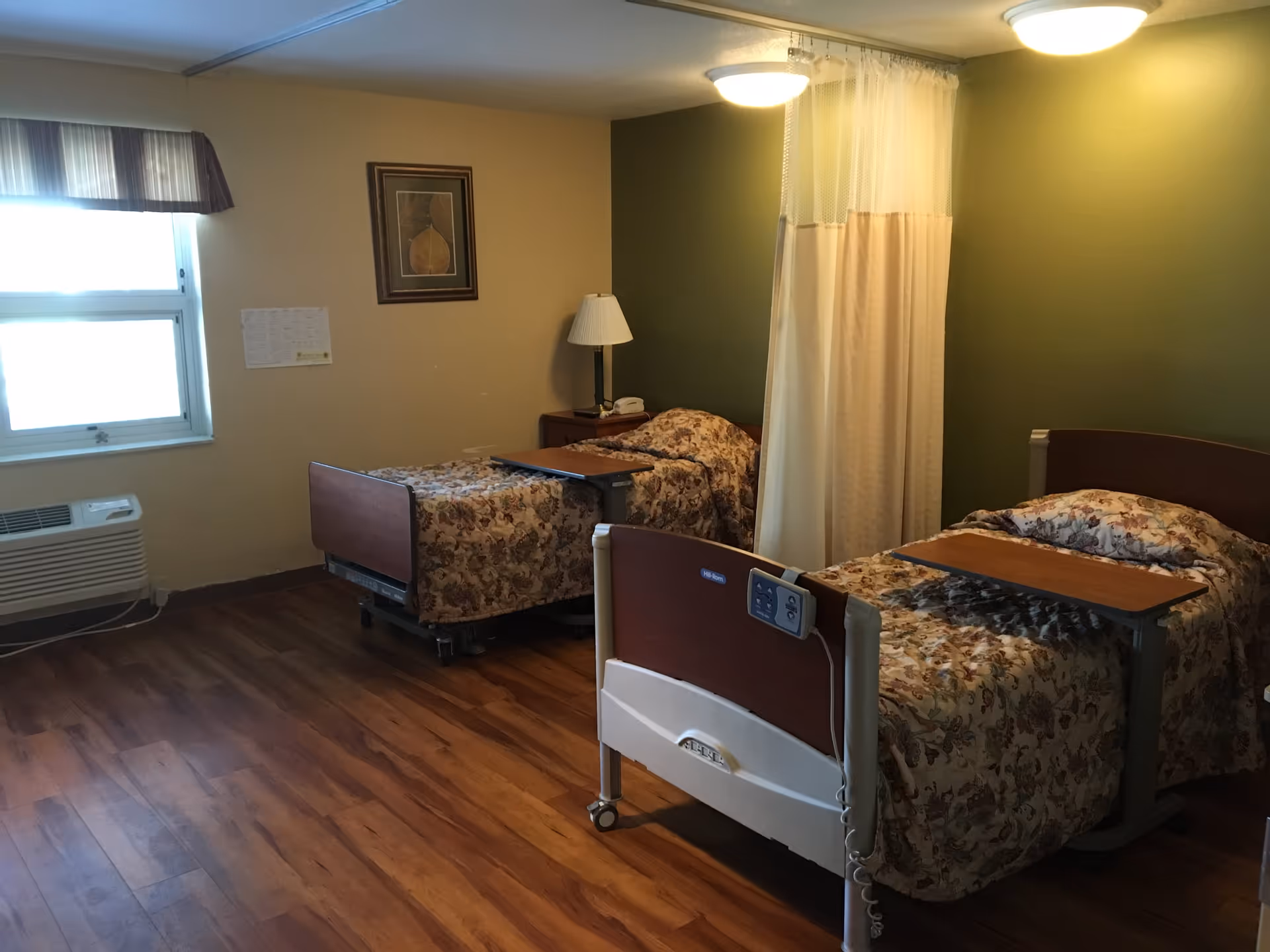 A room with two hospital-style beds, each with floral bedspreads and overbed tables. There is a nightstand with a lamp and a telephone between the beds. A curtain hangs from the ceiling to provide privacy between the beds. The walls are painted in beige and green, and there is a window with a valance on the left side. The floor is wood laminate.