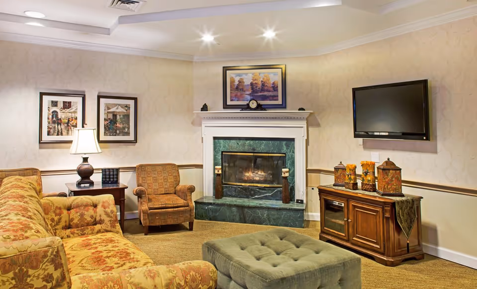 Cozy sitting room with patterned sofas and armchair facing a marble fireplace, wall-mounted TV, and a tufted ottoman.