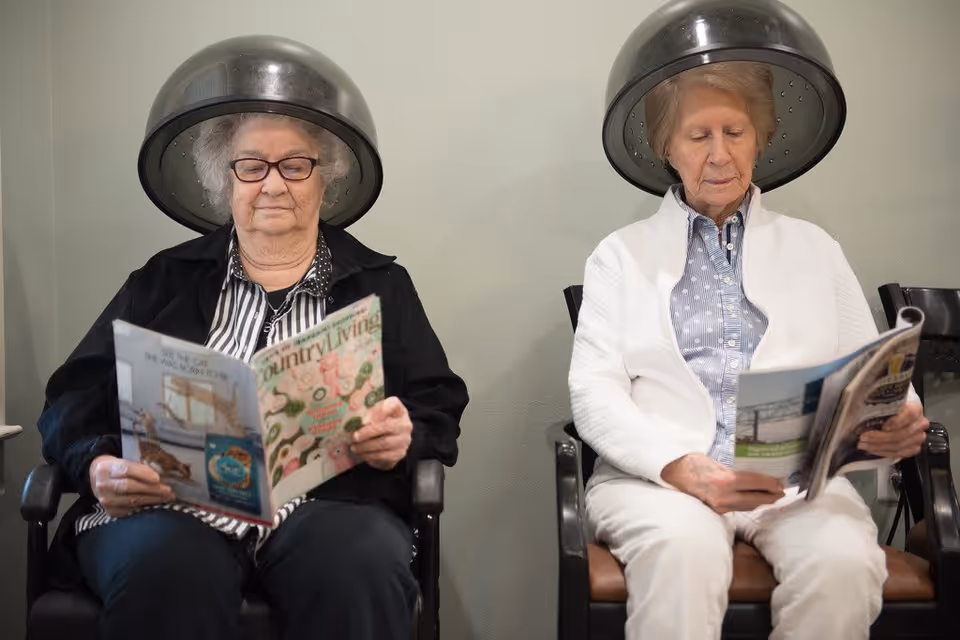 Two elderly women sitting side by side under hair drying hoods, each reading a magazine in a salon-like setting.