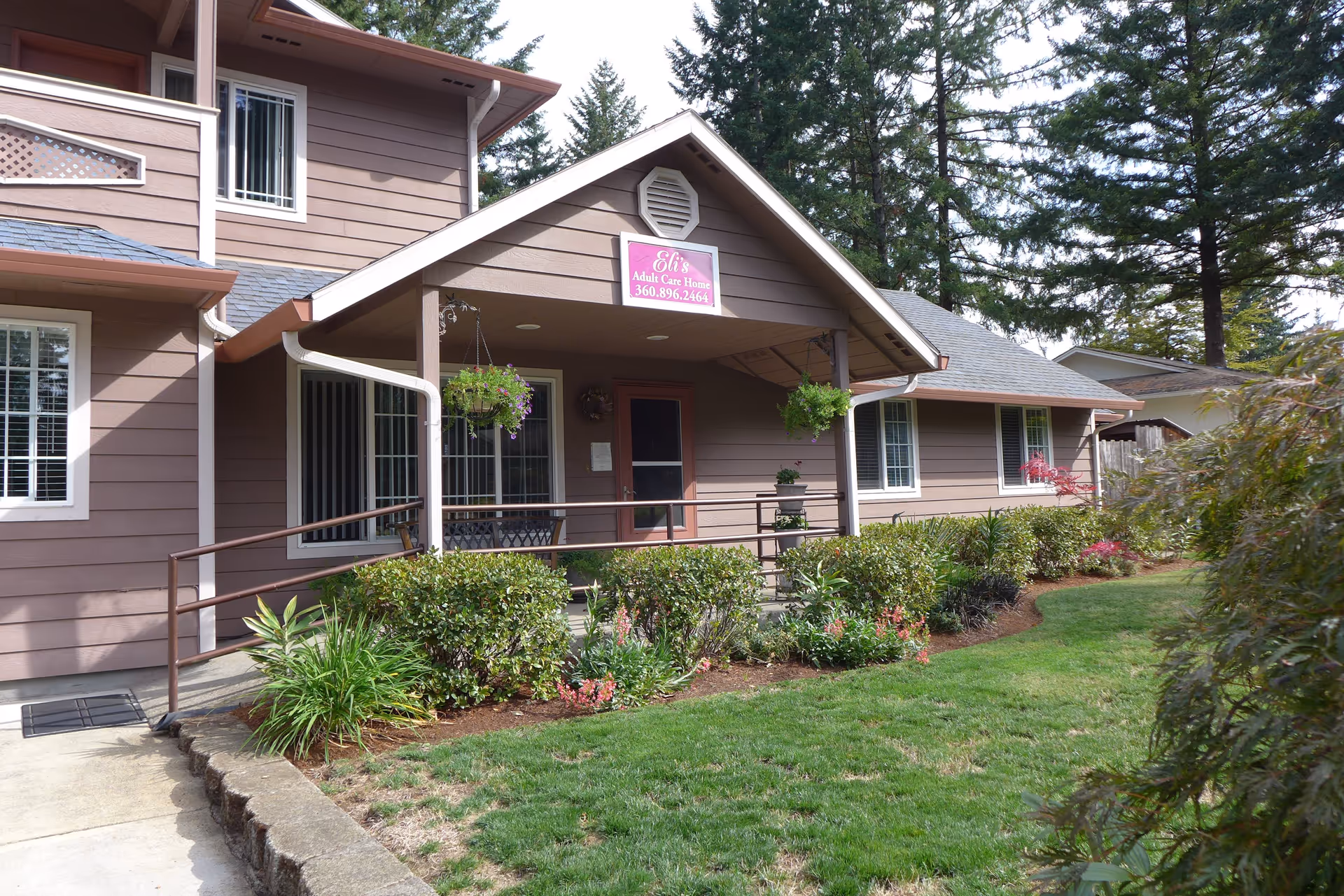 Exterior view of Eli's Adult Care Home, a brown two-story building with a covered porch, hanging flower baskets, and a well-maintained garden with bushes and flowers in front. There is a sign above the porch that reads 'Eli's Adult Care Home 360.896.2464'.