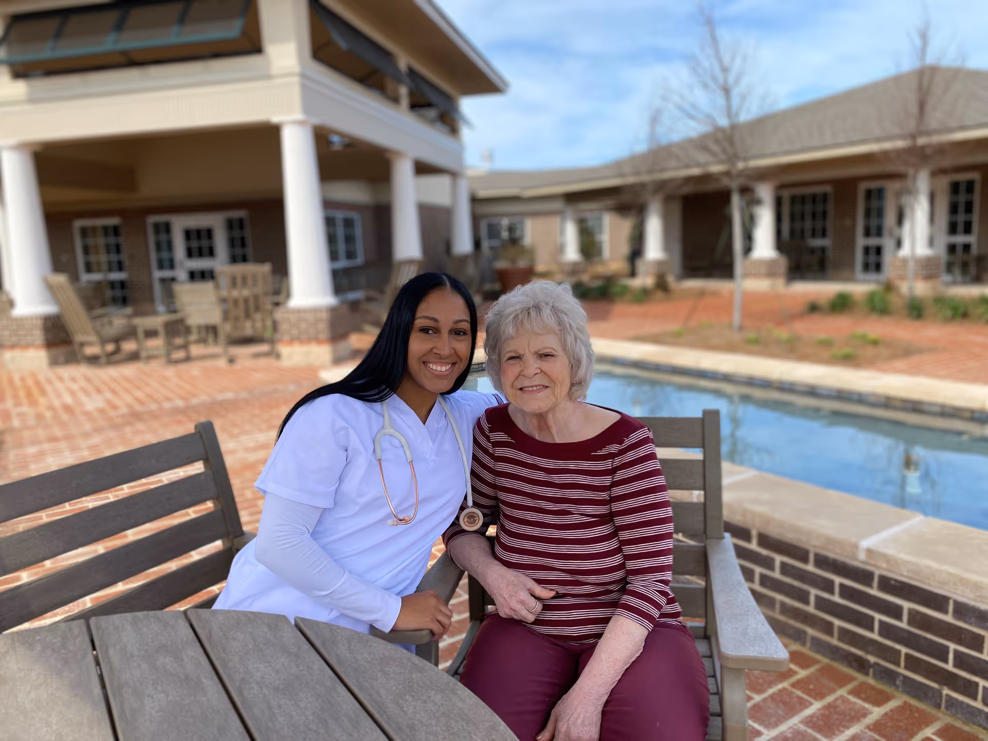 A smiling nurse in white scrubs with a stethoscope around her neck sits next to an elderly woman wearing a red and white striped shirt and maroon pants. They are seated outdoors at a wooden table and chairs near a small pool with a brick patio and building in the background.