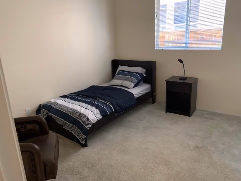 A simple bedroom with a single bed against the wall, made with a striped blue and white comforter and pillows. Next to the bed is a small black nightstand with a modern black lamp on it. There is a window with blinds on the wall above the nightstand, and a brown armchair is partially visible in the corner. The room has beige walls and light-colored carpet flooring.
