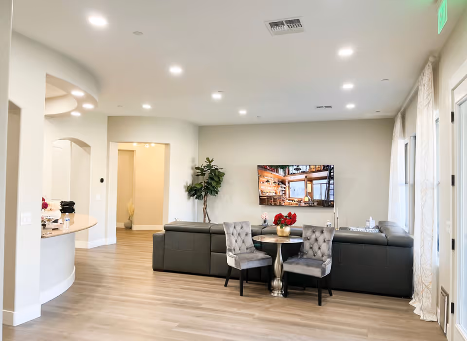 A modern living room area in a senior living facility with a large gray sectional sofa, two gray upholstered chairs around a small round table with a gold vase and red flowers, a flat-screen TV mounted on the wall, light wood flooring, and large windows with light curtains allowing natural light to enter.