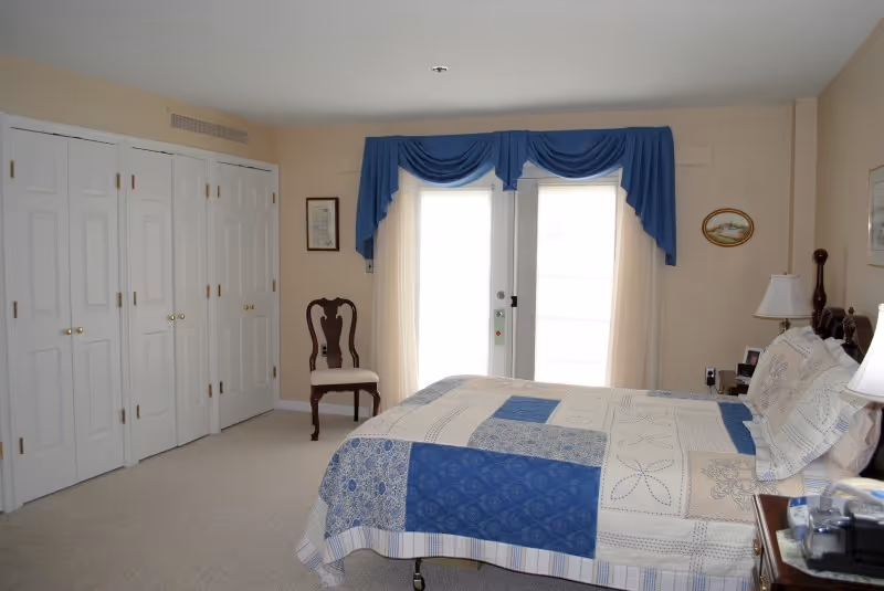 A bedroom with a bed covered in a blue and white patchwork quilt, a wooden chair with a white cushion, white double closet doors, and a glass door with sheer curtains and blue valances.