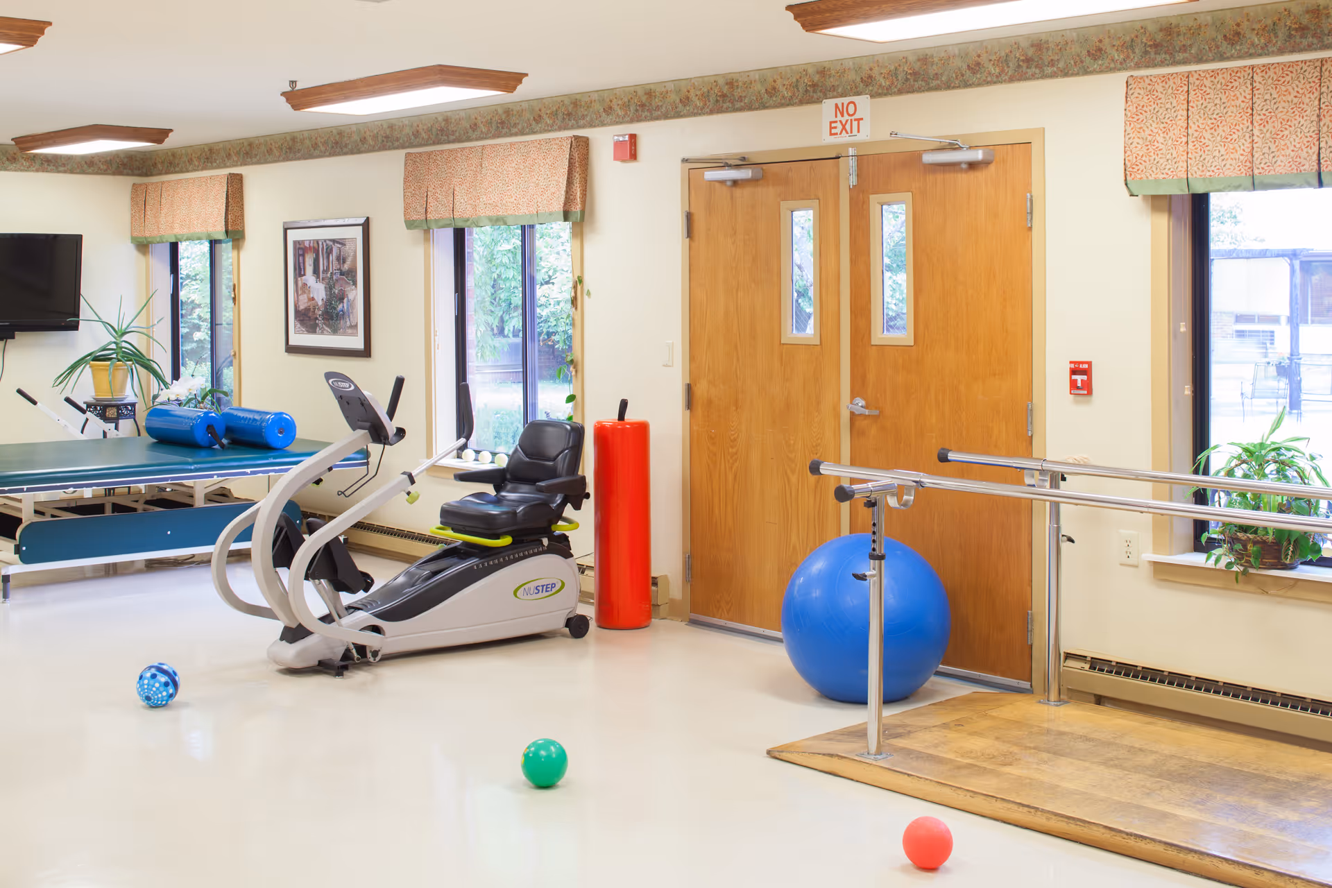 A bright exercise room with various fitness equipment including a NuStep recumbent cross trainer, a ping pong table, a large blue exercise ball, and parallel bars for walking support. The room has large windows with patterned valances, a wall-mounted TV, and a wooden double door with a 'No Exit' sign above it. Several colorful balls are scattered on the floor.