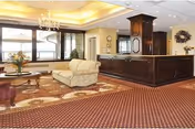 A spacious and well-lit interior room featuring a large patterned carpet, a chandelier, a beige sofa, a wooden coffee table with a flower arrangement, and a dark wooden reception desk or counter. Large windows with blinds allow natural light to enter the room.