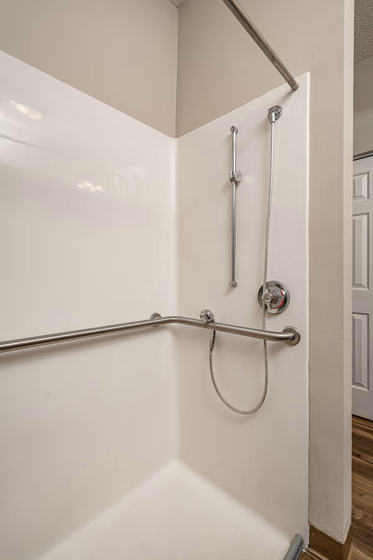 A white shower stall with stainless steel grab bars on the walls and a handheld showerhead attached to a vertical sliding bar. The shower control knob is visible, and the floor outside the shower has wood-style flooring.