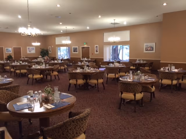 A spacious dining room with multiple round tables set with placemats, glasses, and silverware. The room has patterned carpet, beige walls with framed artwork, large windows letting in natural light, and chandeliers hanging from the ceiling.