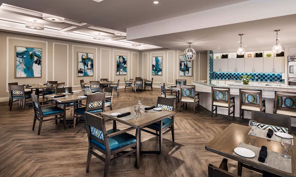 A spacious dining room with multiple wooden tables and chairs arranged neatly. Each table is set with plates, glasses, and black napkins. The room features a modern design with abstract blue and green paintings on the walls, a wooden floor with a herringbone pattern, and a kitchen area with a blue tiled backsplash and bar seating with high chairs. Ceiling lights and pendant lamps provide illumination.