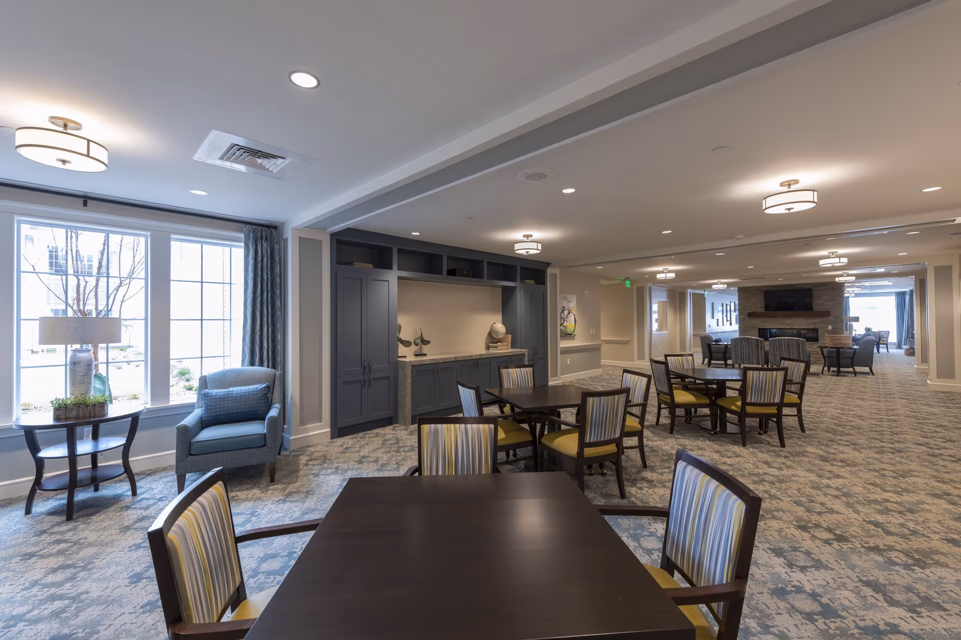 A spacious and well-lit common area in a senior living facility featuring multiple dark wood tables with striped cushioned chairs, a cozy seating area with armchairs near large windows, built-in cabinetry, and a stone fireplace with a mounted TV in the background.