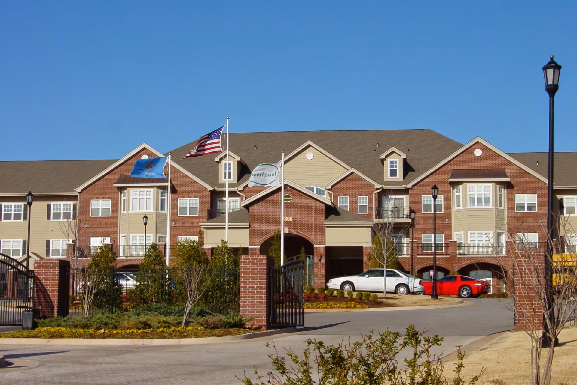 Exterior view of a senior living facility building with brick and beige siding, multiple windows, and three flagpoles displaying the American flag and other flags. There is a gated entrance with brick pillars and a driveway with parked cars under a clear blue sky.