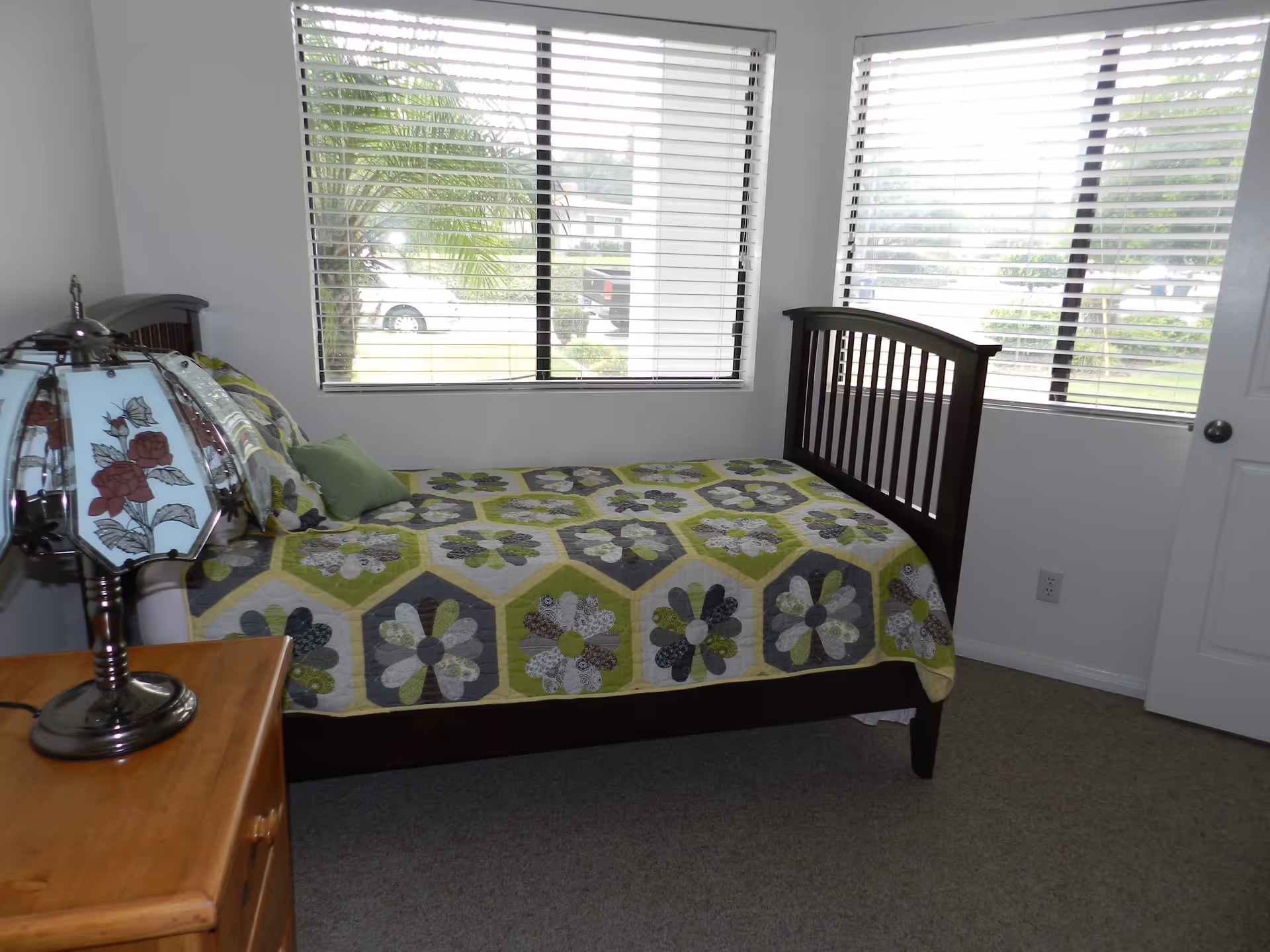 Small bedroom with a wooden single bed covered by a green and gray floral quilt, windows with blinds, and a bedside table with a decorative lamp.