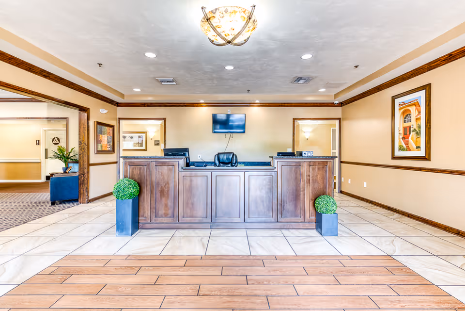 Well-lit senior living lobby with a central wooden reception desk, two potted ornamental shrubs, framed artwork, and a wall-mounted TV.