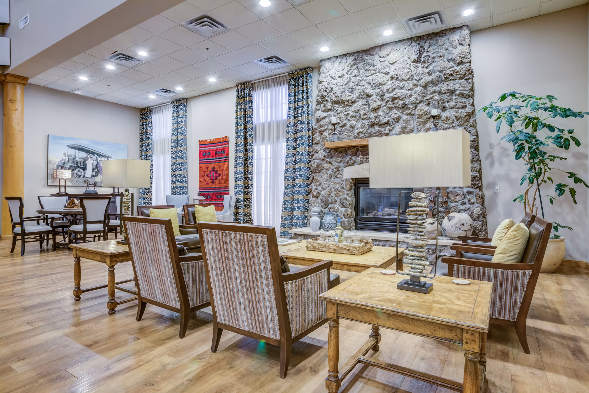 A cozy senior living community lounge area featuring wooden chairs with striped cushions, a stone fireplace, wooden tables with lamps, tall windows with patterned curtains, a large potted plant, and a dining table with chairs in the background.