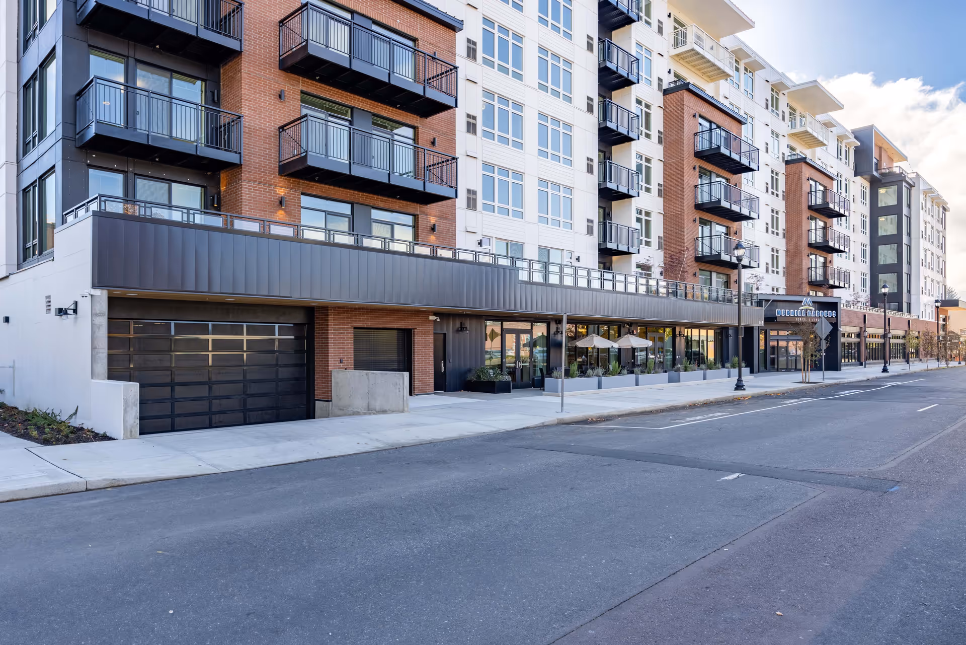 Exterior view of a modern multi-story residential building with balconies, large windows, and a street in front. The building has a mix of brick and light-colored siding with a sign that reads Merrill Gardens Senior Living. There are outdoor seating areas with umbrellas near the entrance.