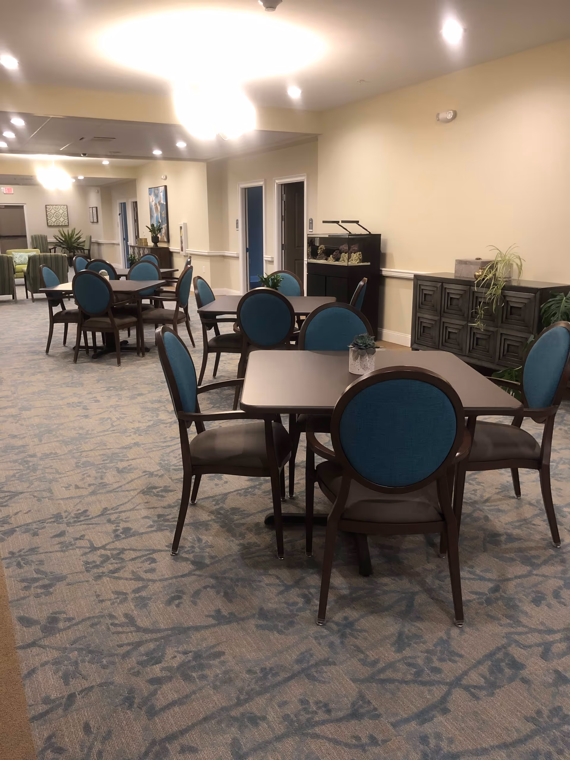 Interior view of a senior living facility dining area with multiple square tables and chairs featuring blue backs and brown seats. The room has beige walls, patterned carpet, and ceiling lights. There is a fish tank and decorative plants on a cabinet against the wall.