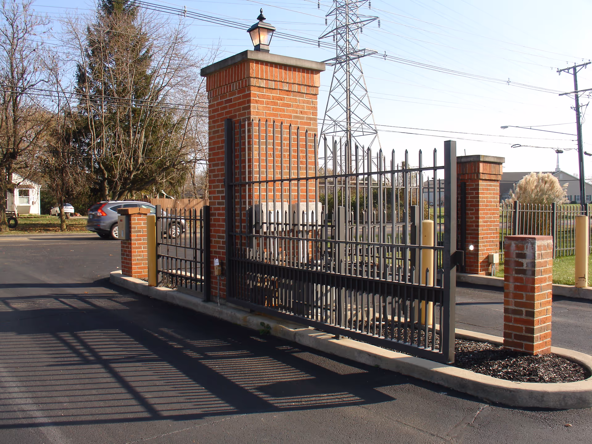 Brick-pillared gated entrance with a metal security gate casting long shadows on the asphalt driveway.