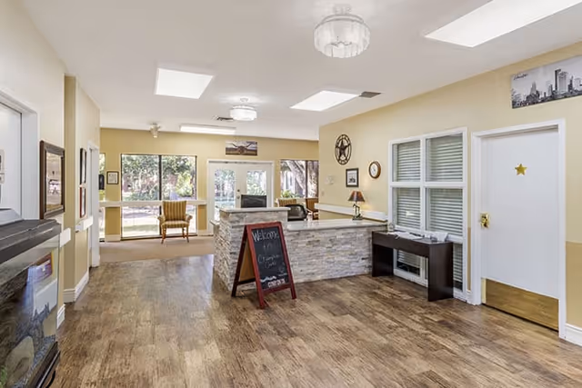 Interior view of a bright and welcoming assisted living community common area with wood flooring, a stone-faced reception desk with a chalkboard sign, seating area with chairs near large windows, and light yellow walls decorated with framed pictures and a clock.
