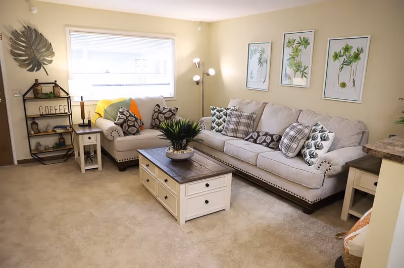 A cozy living room with a beige carpet and light yellow walls. There is a large beige sofa and a matching loveseat adorned with various patterned throw pillows. A wooden coffee table with drawers sits in front of the sofa, topped with a green potted plant. On the wall above the sofa are three framed botanical prints. To the left, a small shelving unit labeled 'COFFEE' holds decorative items. A floor lamp with multiple lights stands in the corner near a window with blinds partially open, allowing natural light to fill the room.
