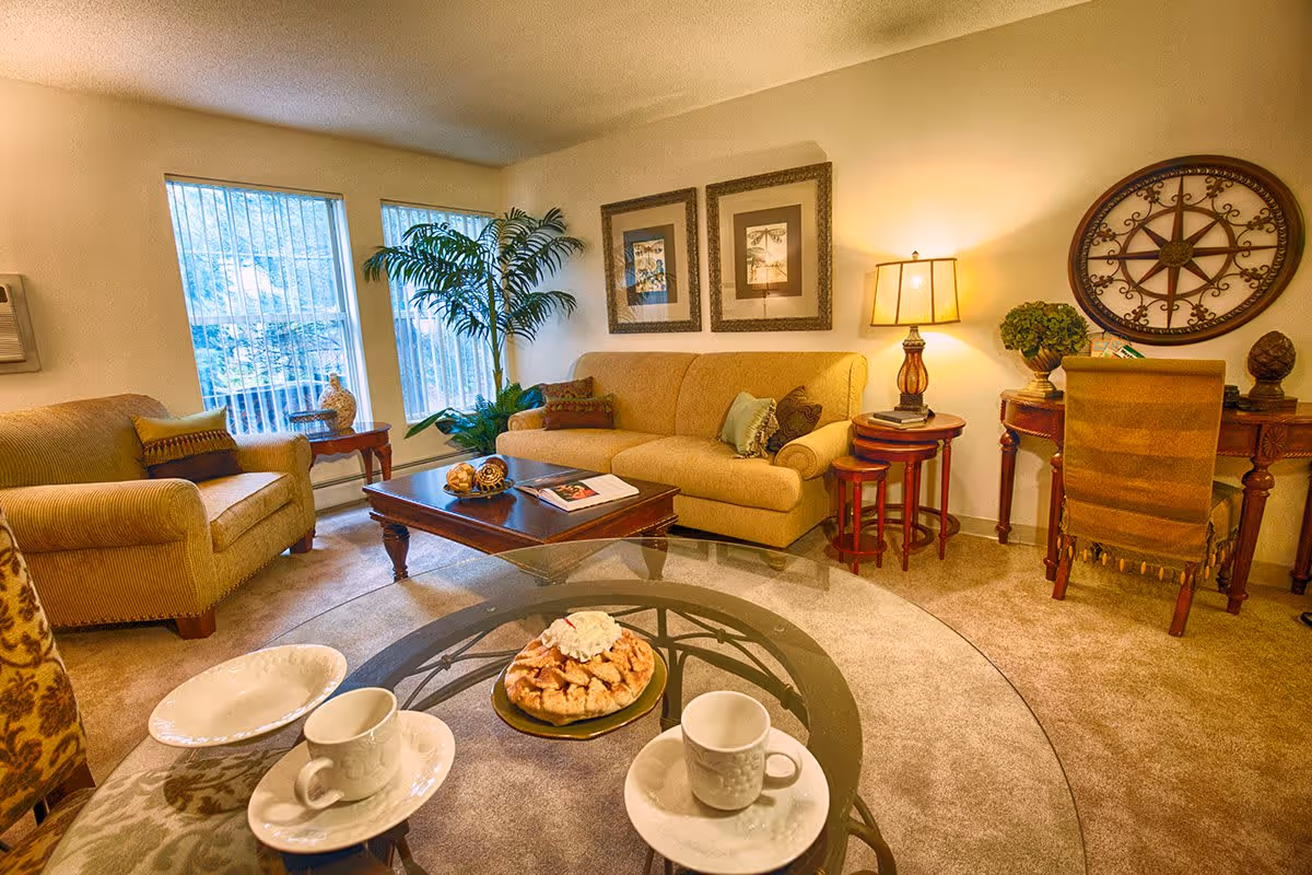 Bright, warmly lit living room with a sofa and armchairs around a coffee table, decorative wall art, and a glass table set with cups and a pastry.