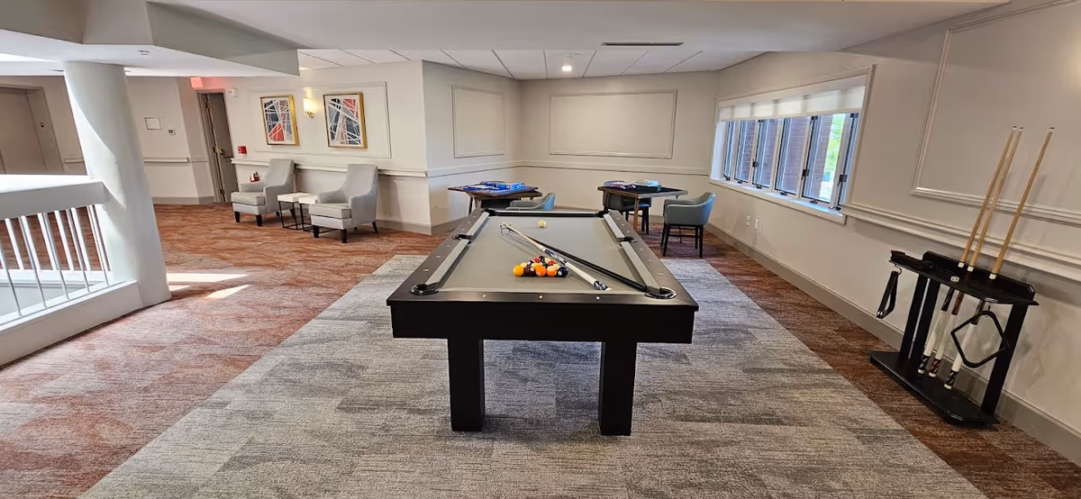 A recreational room in Riverton Enhanced Senior Living featuring a pool table with balls and cues, two armchairs with a small table against the wall, two small tables with chairs near a window, and a rack holding pool cues. The room has carpeted flooring and neutral-colored walls with framed abstract artwork.