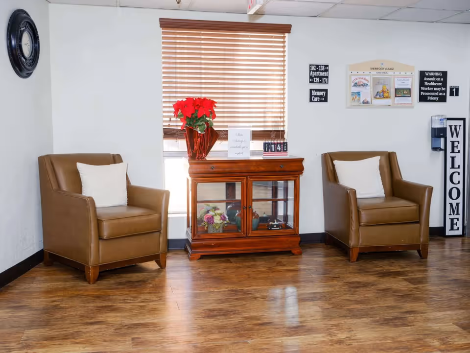 A cozy seating area in Sherwood Village Assisted Living & Memory Care featuring two brown leather armchairs with white cushions, a wooden cabinet with glass doors displaying decorative items, a window with wooden blinds, a red poinsettia plant on top of the cabinet, and various signs on the wall including a large vertical 'WELCOME' sign and informational plaques.