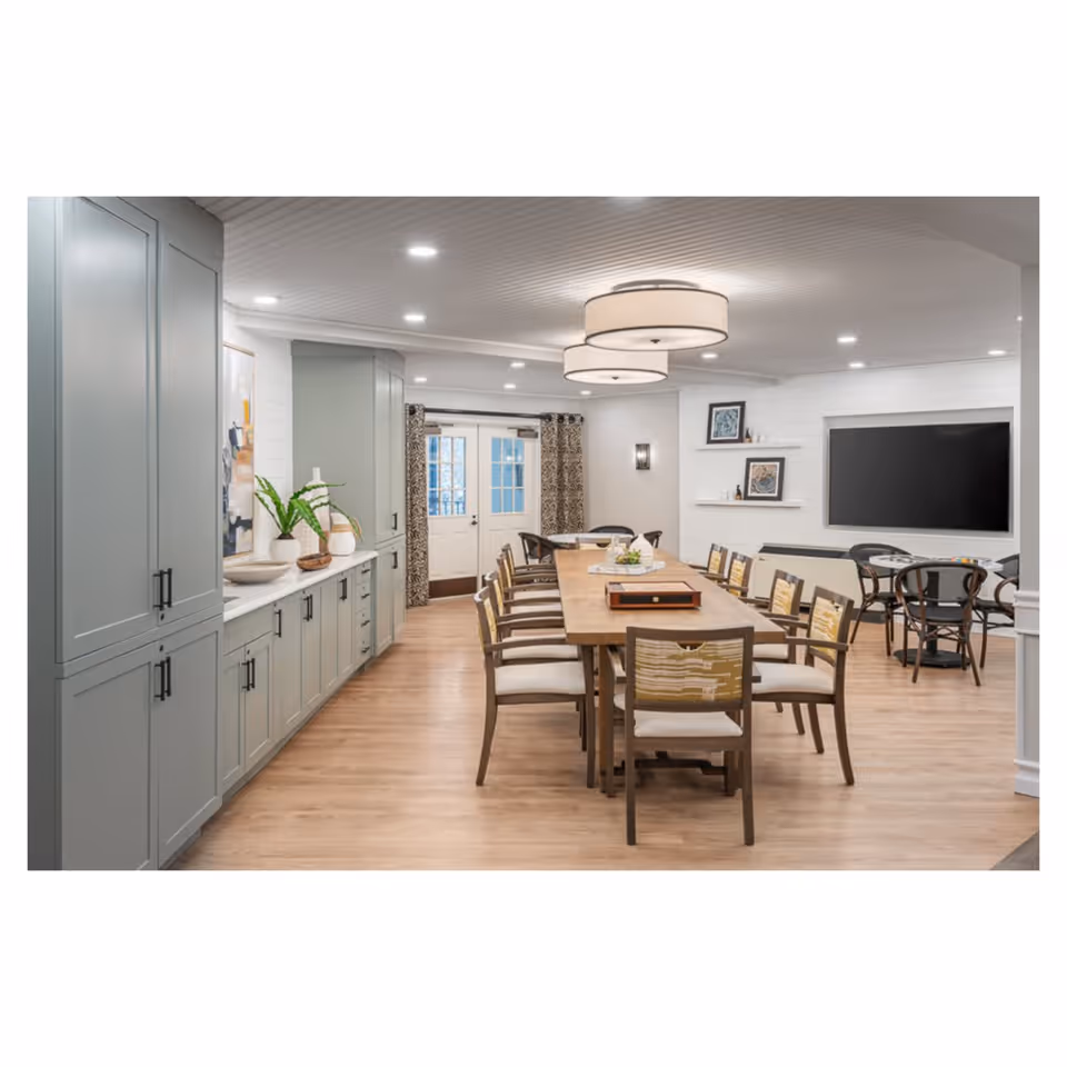A spacious, well-lit dining room with a long wooden table surrounded by chairs. The room features light wood flooring, gray cabinetry along one wall with decorative items, two large circular ceiling lights, a flat-screen TV mounted on the wall, and additional smaller tables and chairs in the background near a set of double doors with glass panes and patterned curtains.