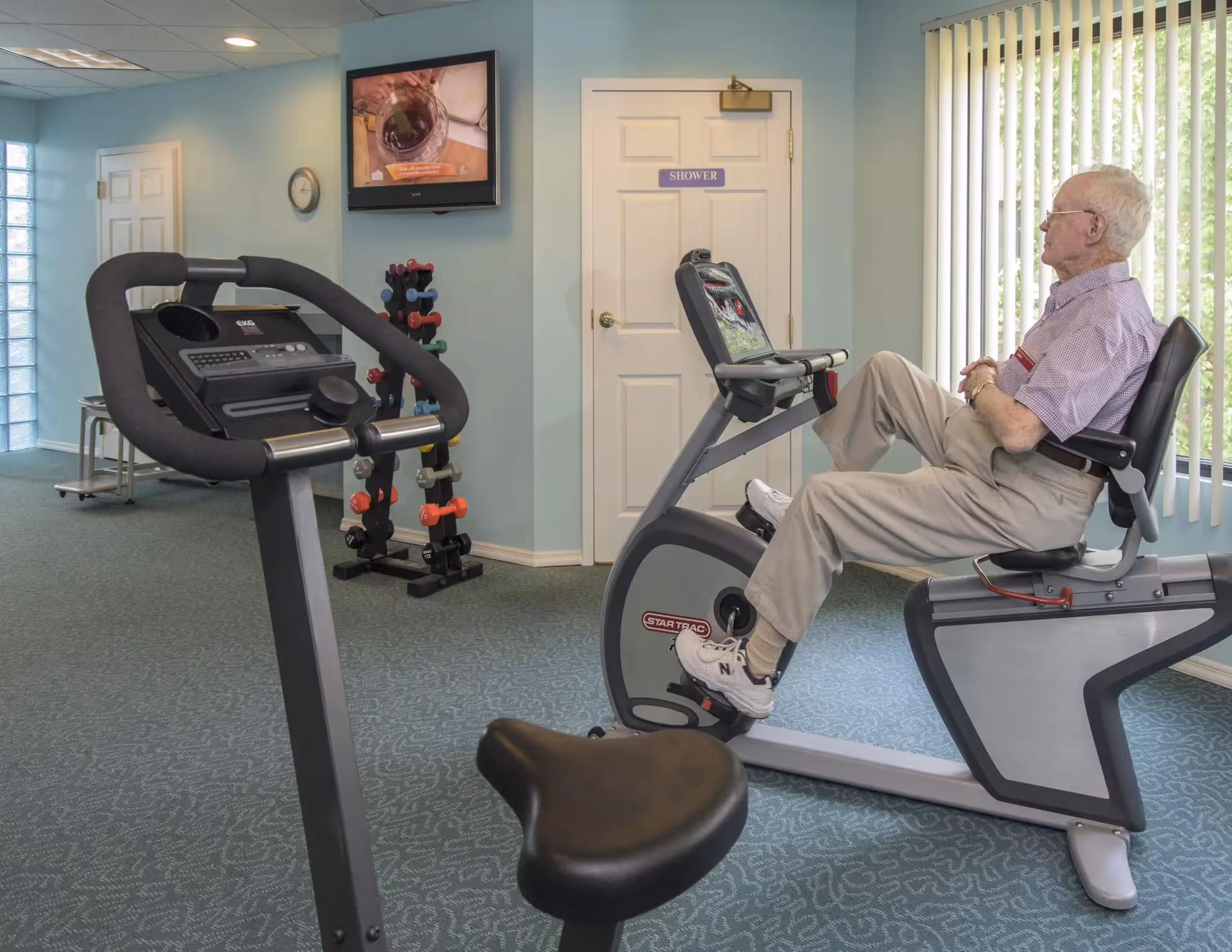 An elderly man is seated on a recumbent exercise bike in a fitness room with light blue walls and carpeted floor. There is another exercise bike in the foreground, a rack of colorful dumbbells against the wall, a wall-mounted TV showing a cooking program, and a door labeled 'SHOWER'. Vertical blinds cover a large window letting in natural light.