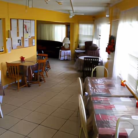 Interior view of a senior living facility common area with yellow walls, multiple tables covered with tablecloths, chairs, a bulletin board on the wall, and a seating area with armchairs and a lamp near windows with sheer curtains.