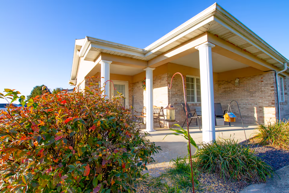 Front porch area of a single-story brick building with white columns supporting the roof overhang. There are chairs and a bench on the porch, along with bird feeders and some plants and bushes in the foreground under a clear blue sky.