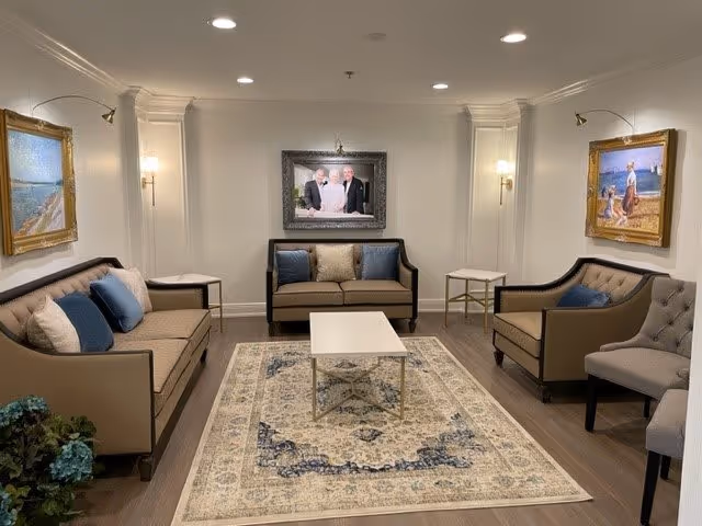 A cozy living room with beige walls and wooden flooring featuring two beige sofas with blue and beige cushions, a beige armchair, a white coffee table on a patterned rug, and framed paintings on the walls. The room is softly lit with recessed ceiling lights and wall sconces.