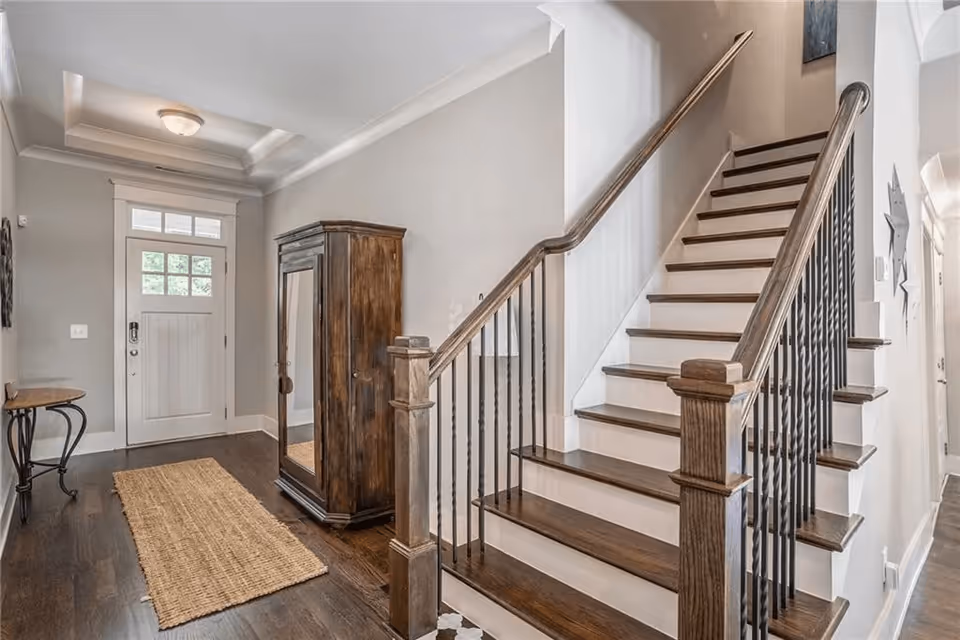 Interior view of a hallway featuring a wooden staircase with dark handrails and white risers on the right side. A wooden armoire with a mirrored door stands against the wall, and a small table with curved legs is positioned near the front door. The floor is dark hardwood with a woven rug runner, and the walls are painted light gray. The front door has a window with multiple panes at the top.