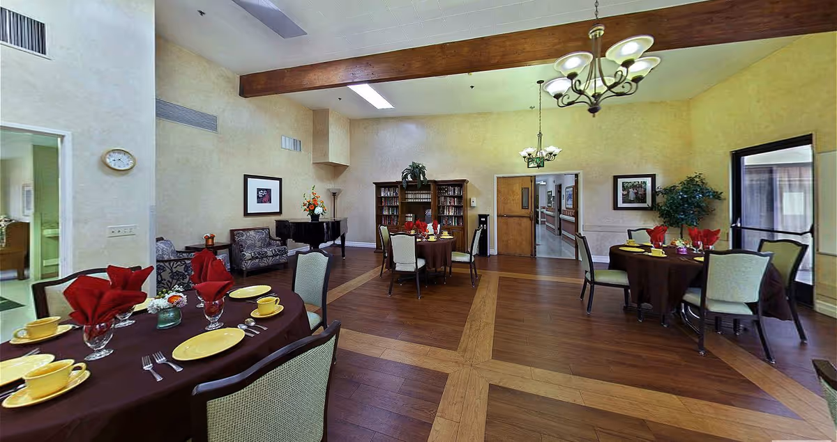 A dining room in a senior living facility with round tables covered in dark brown tablecloths, set with yellow plates, cups, and red folded napkins. The room has wooden flooring with a patterned inlay, beige walls, and ceiling lights with multiple bulbs. There is a bookshelf filled with books, a piano, framed pictures on the walls, and a potted plant near a glass door leading outside. A hallway is visible through a door in the back.