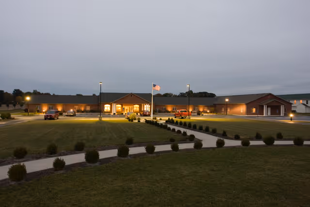 Exterior view of Putnam Heritage Assisted Living facility at dusk with lights on, a flagpole with an American flag in front, a parking lot with several vehicles, and a landscaped lawn with small bushes and paved walkways.