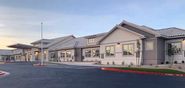 Exterior view of a single-story senior living facility building with a tiled roof, multiple windows, and a covered entrance. The surrounding area includes a paved driveway and landscaped areas with small plants and trees under a clear sky at sunset.