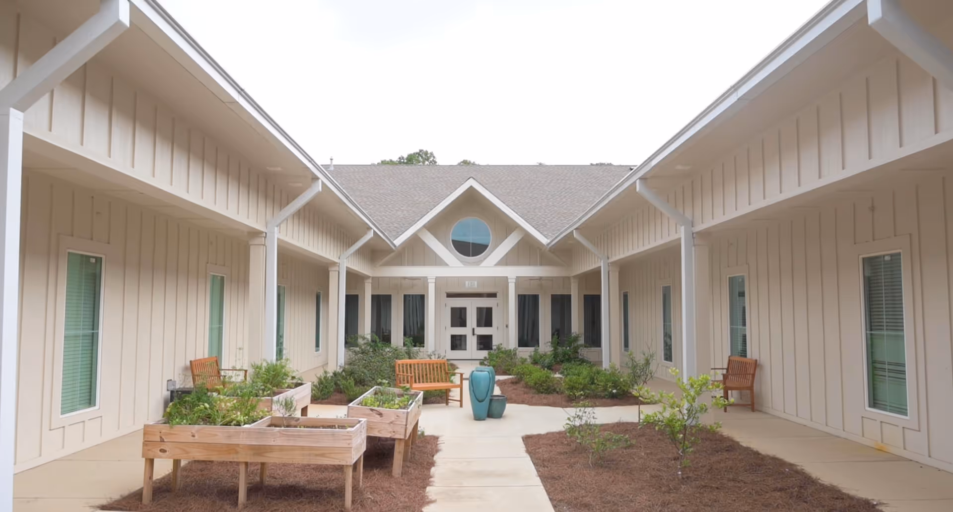 Courtyard between two wings of a beige building with raised garden beds, benches, and a paved walkway leading to a central entrance.