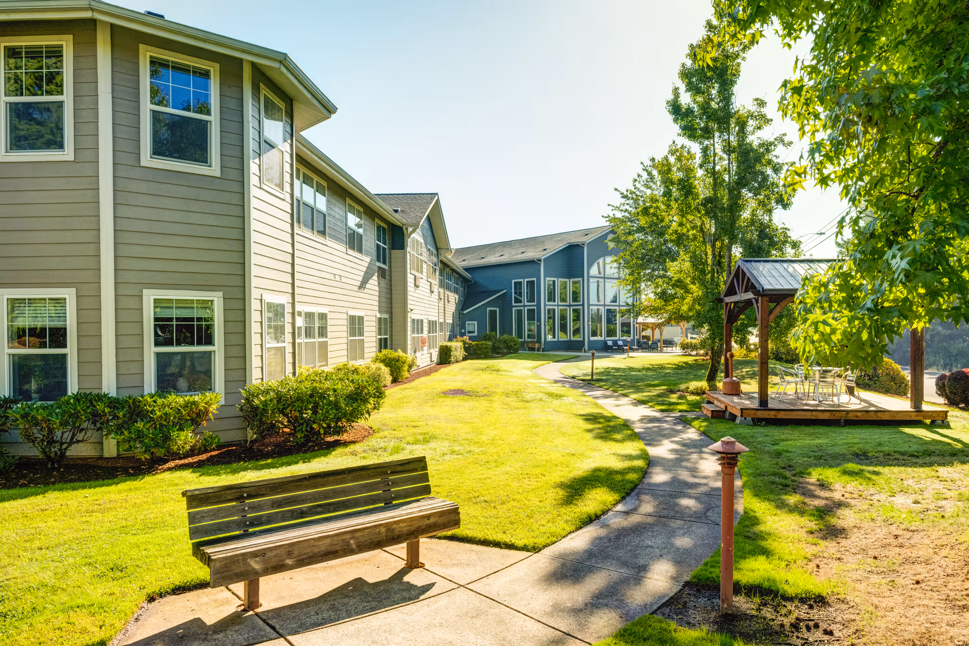 Outdoor view of Callahan Village Retirement and Assisted Living showing a paved walkway curving through a grassy area with a wooden bench on the left and a small gazebo with chairs and tables on the right. The building has multiple windows and is surrounded by trees and shrubs under a clear sky.