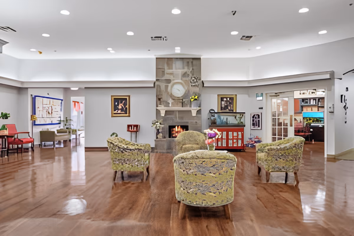 Spacious senior living lounge with upholstered chairs arranged around a central stone fireplace and polished wood floors.