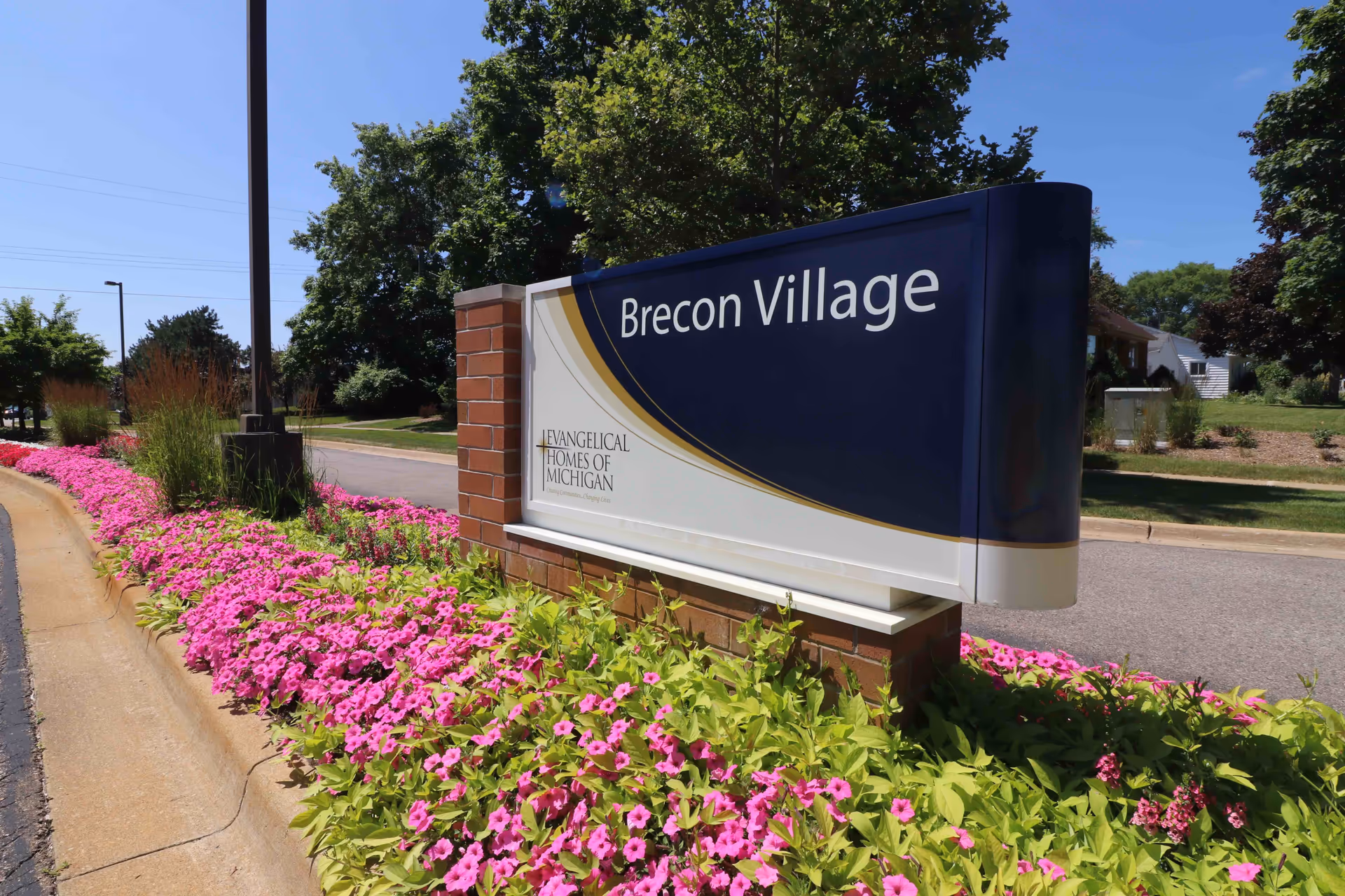 Entrance sign reading "Brecon Village" beside a flowerbed of pink flowers and a roadway with trees in the background.