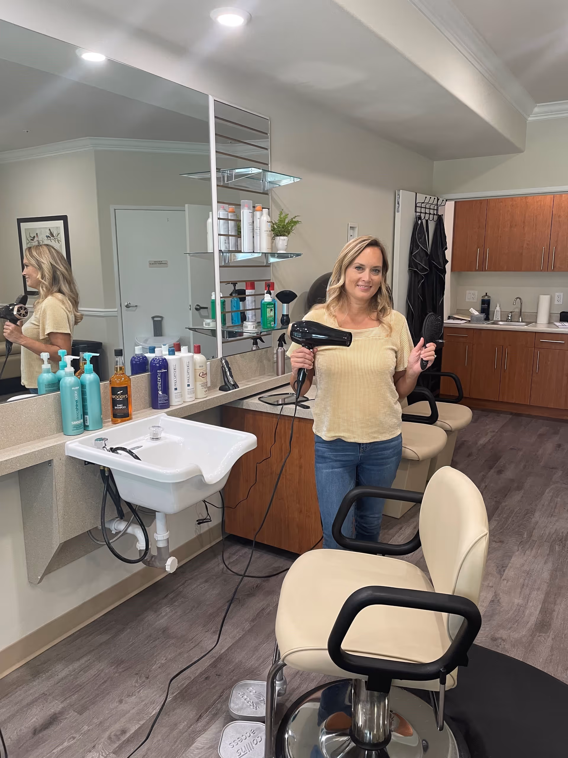 A woman stands in a salon area holding a hairdryer and brush between styling chairs, sinks, mirrors, and hair products.