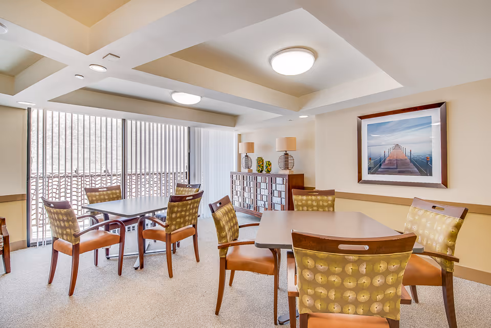 Bright common dining room with several tables, upholstered chairs, a sideboard, and framed artwork on the wall.