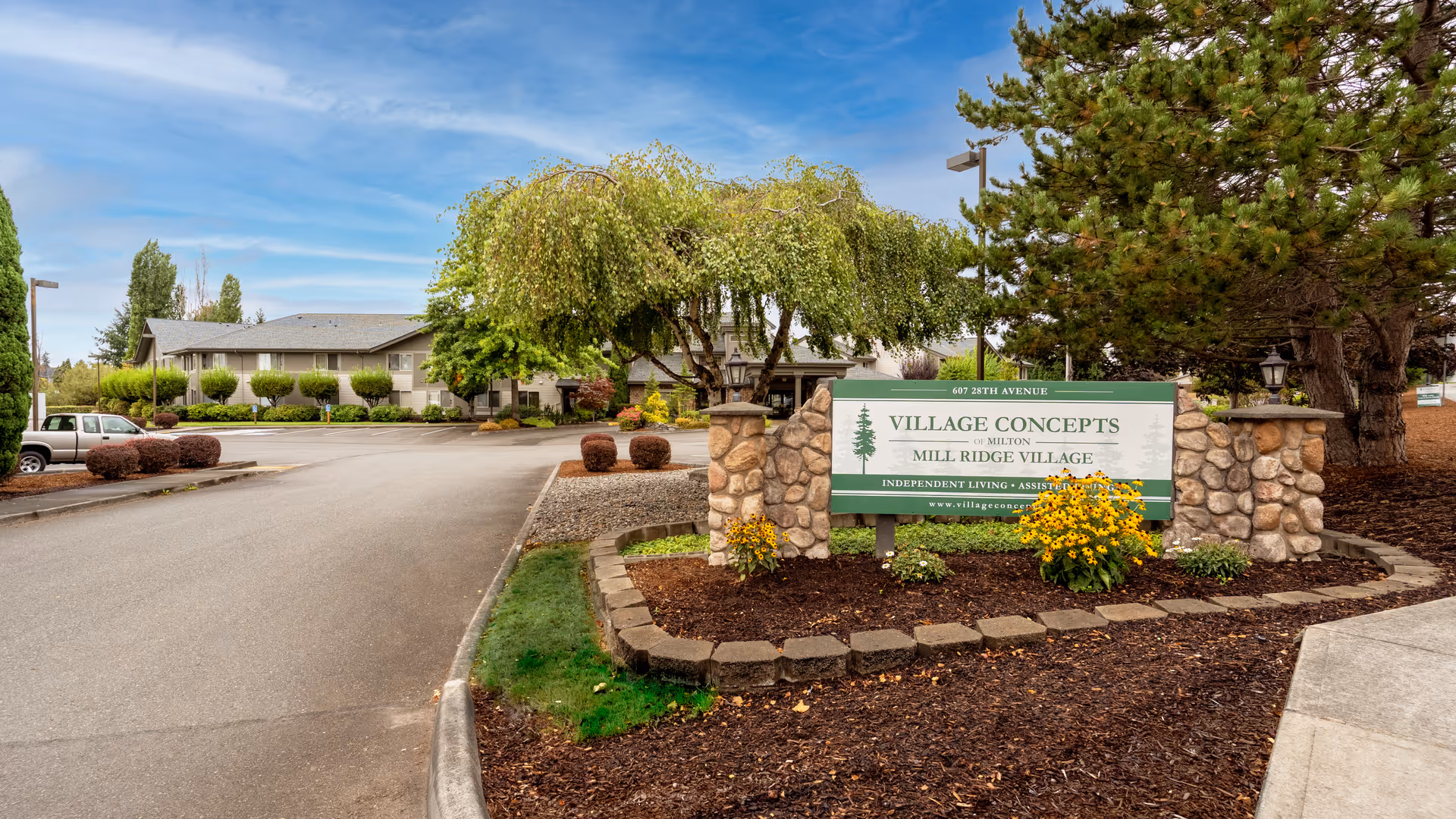 Entrance area of Village Concepts of Milton - Mill Ridge Village senior living facility with a stone-bordered flower bed featuring yellow flowers and a sign displaying the facility name. The building and parking area are visible in the background under a partly cloudy blue sky.