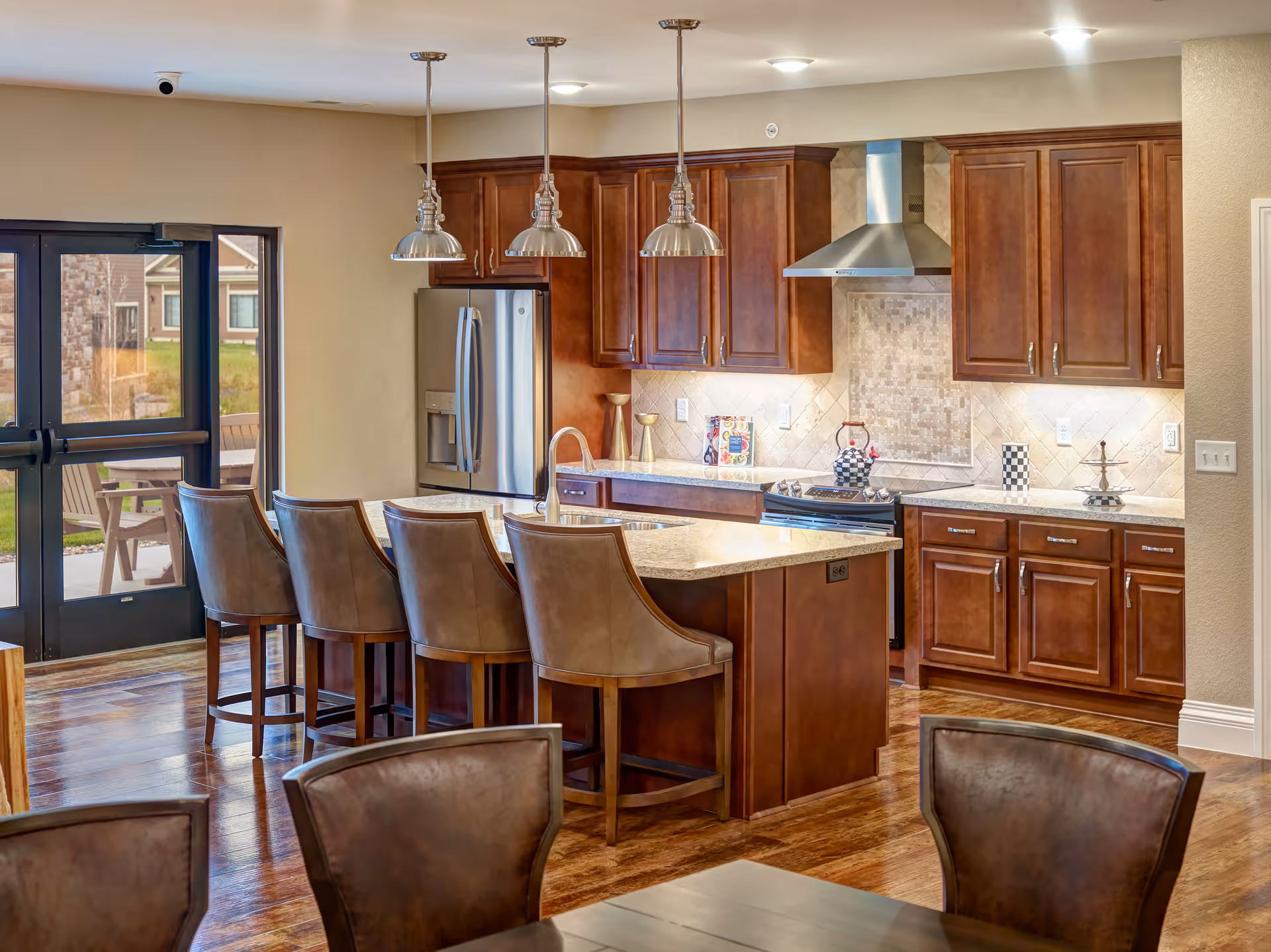 A modern kitchen with wooden cabinets, a large island with a granite countertop, and four brown leather bar stools. Stainless steel refrigerator and stove with a vent hood are visible. Three pendant lights hang above the island. There is a glass door leading outside, and wooden flooring throughout.