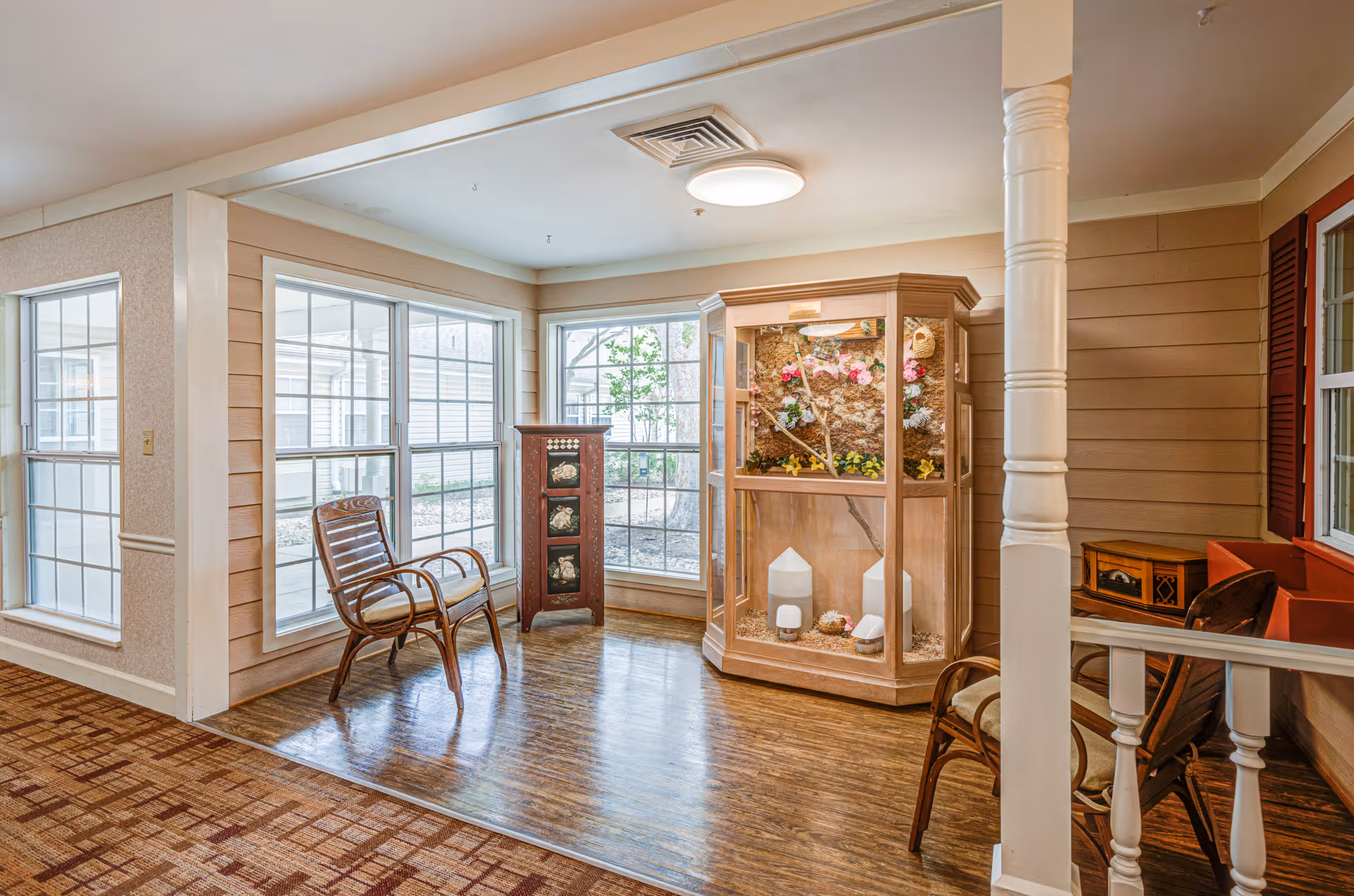 Bright seating nook with wicker chairs, a decorative display cabinet, and large windows in a memory care facility.